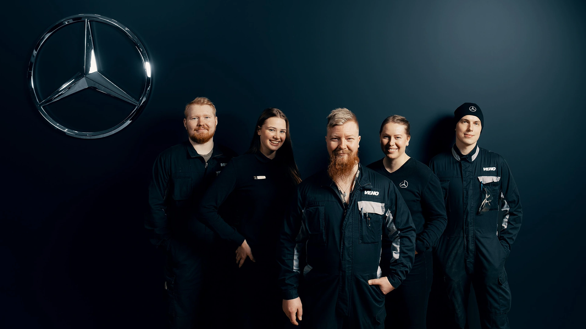Veho servisa darbinieki stāv pie tumšas sienas Veho servisa centrā