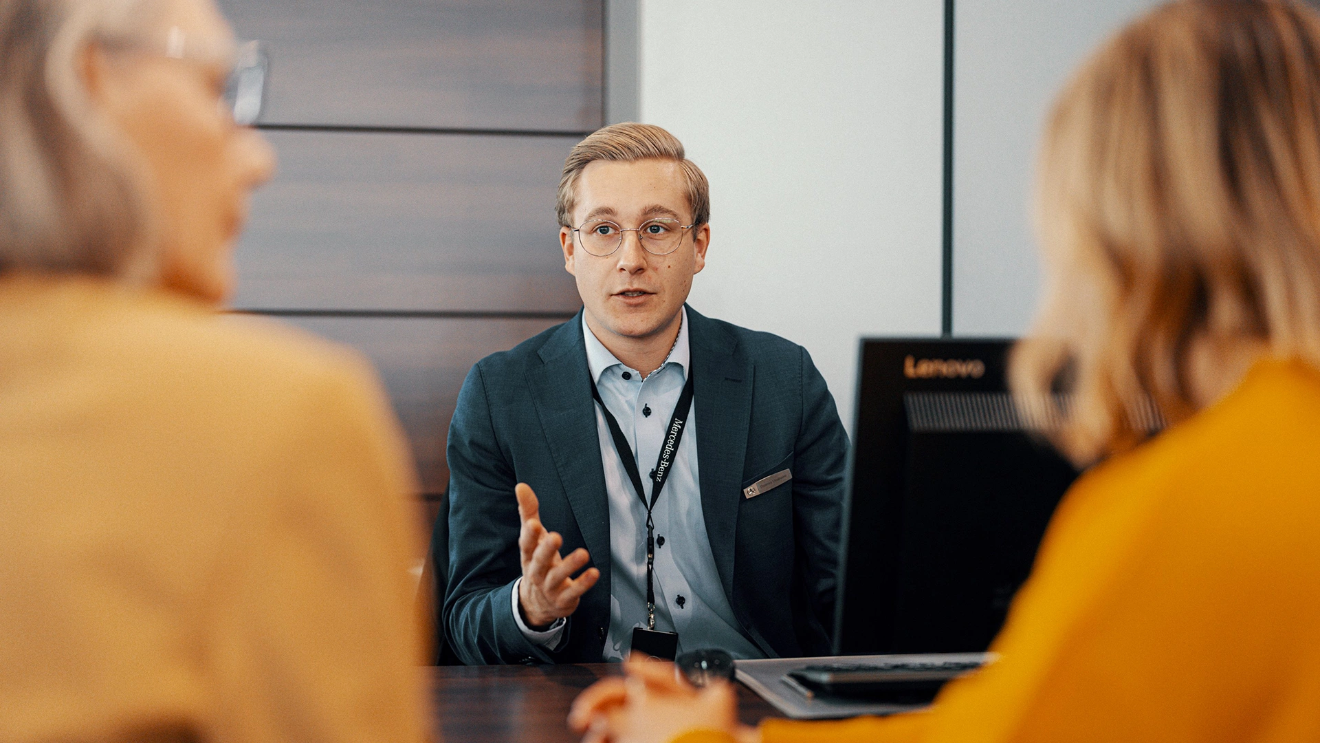 Automašīnu pārdevējs sēž pie rakstāmgalda un sarunājas ar diviem klientiem.